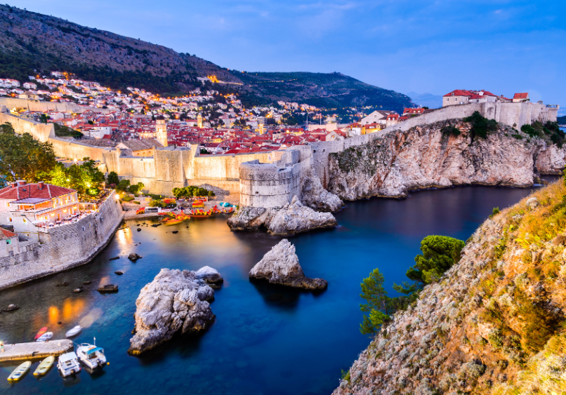Aerial view of Dubrovnik’s old town and stone city walls at dusk, with rocky coastline, calm blue water, and boats docked near the shore.