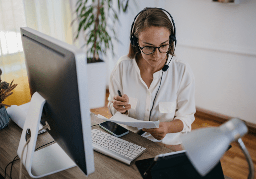 A woman wearing a headset sits at a desk with a computer, taking notes on a piece of paper while working from home.