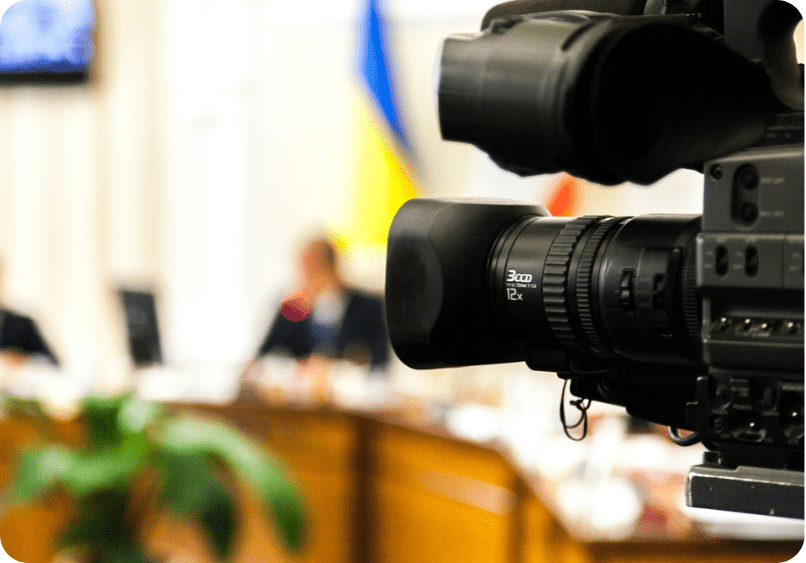 Close-up of a professional video camera recording a meeting in a conference room, with blurred people and a flag in the background.