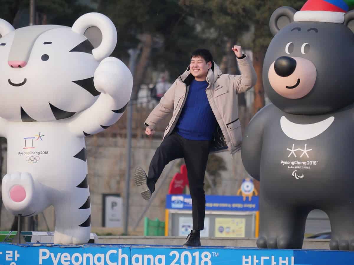 A person poses playfully between two large PyeongChang 2018 Olympic mascots, Soohorang the white tiger and Bandabi the black bear, outdoors.
