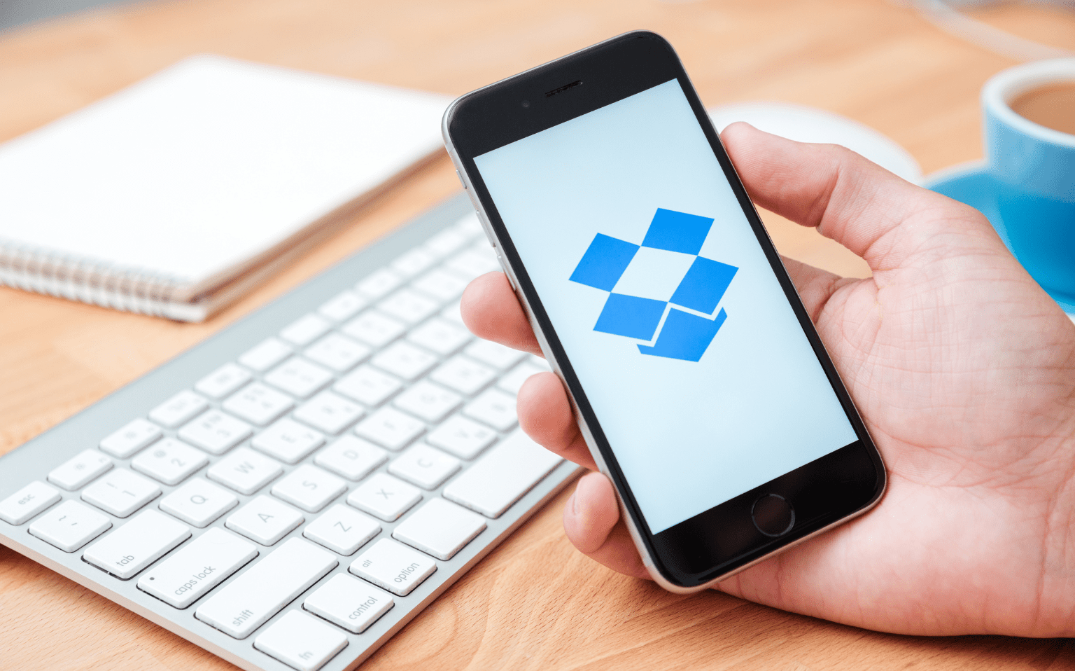 A person holds a smartphone displaying the Dropbox logo, with a keyboard, notepad, and coffee cup on a wooden desk in the background.