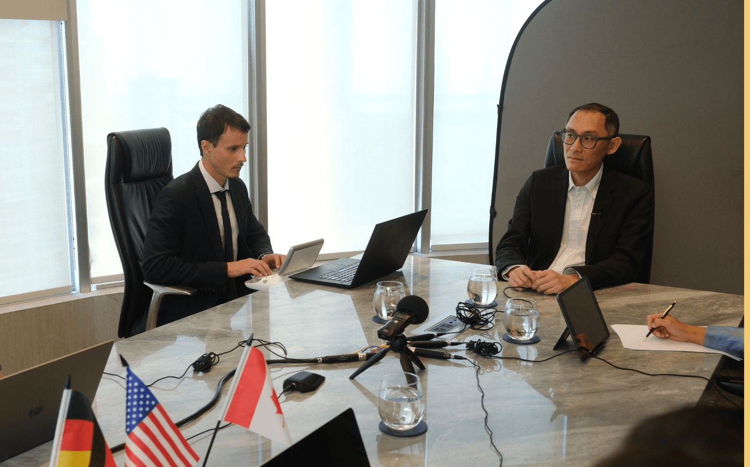 Two men sit at a conference table with laptops, microphones, and country flags. One is typing while the other listens. Glasses of water and notepads are also on the table.