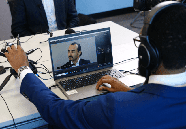 A person wearing headphones edits a video of a man speaking, displayed on a laptop screen, at a desk with microphones and recording equipment.