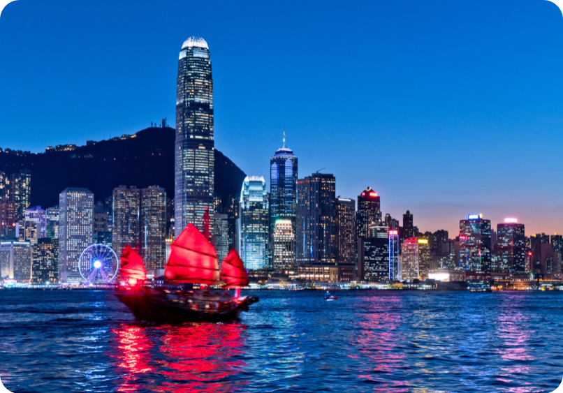 A traditional junk boat with red sails glides across Victoria Harbour at dusk, with the illuminated Hong Kong skyline in the background.