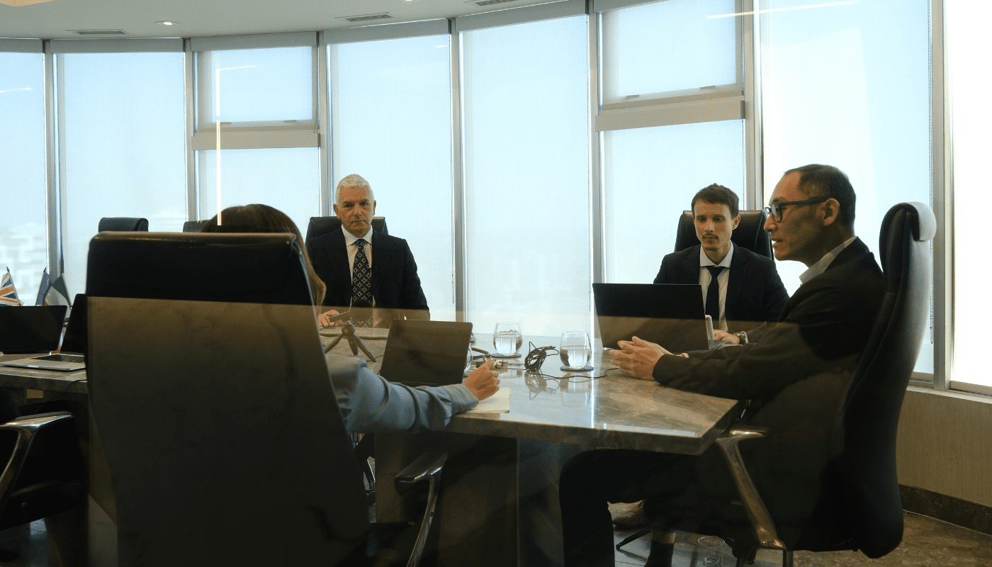 Four people in business attire sit around a conference table in a modern office, engaged in a meeting with laptops and documents in front of them.