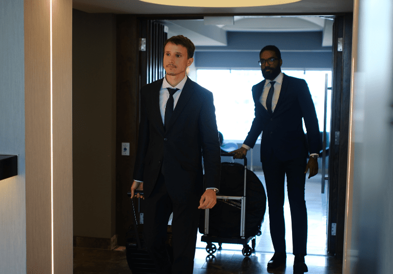 Two men in business suits enter a room; one carries a briefcase, the other pulls a suitcase.