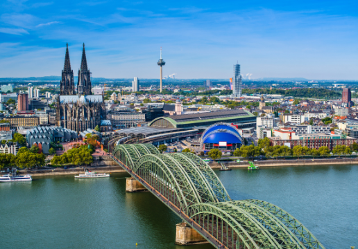Aerial view of Cologne showing the Hohenzollern Bridge crossing the Rhine River, Cologne Cathedral, and city buildings under a clear blue sky.
