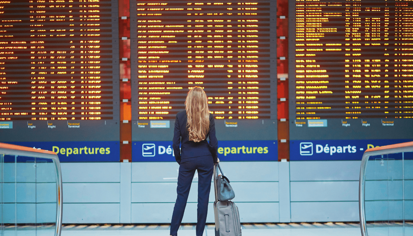 A person with long hair stands with a suitcase, looking at a large departures board displaying flight information at an airport.