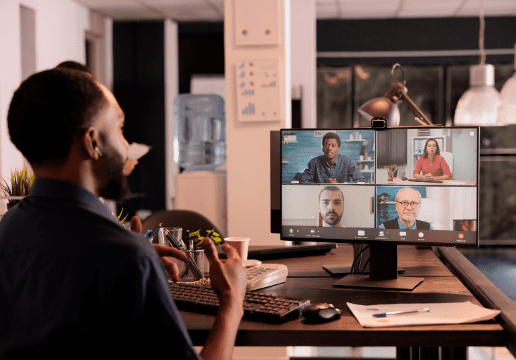 A person sits at a desk in an office, participating in a video conference with four people shown on a computer monitor.