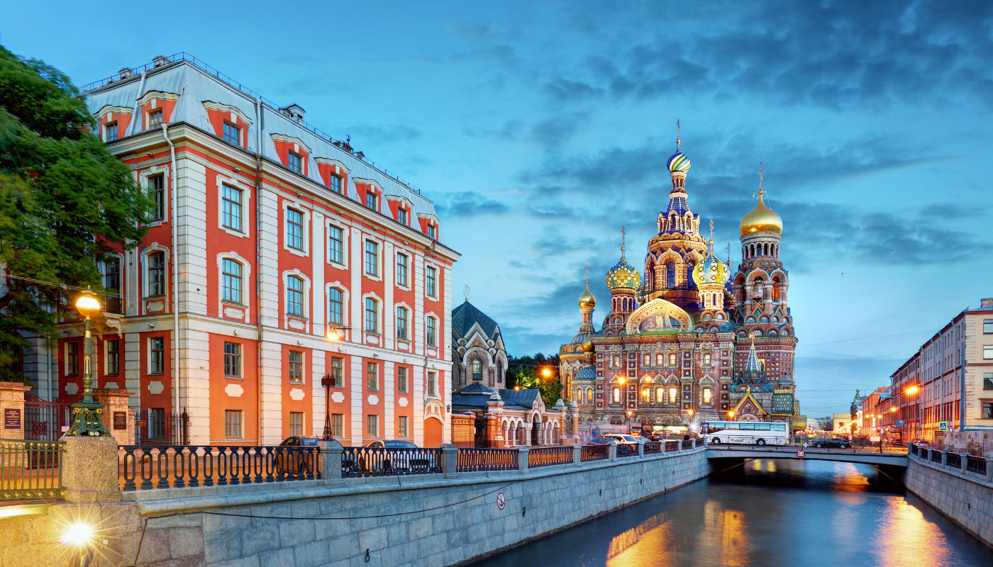 A view of the Church of the Savior on Spilled Blood and a historic building along a canal in St. Petersburg, Russia, at dusk.