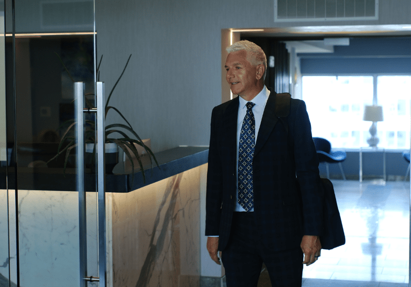 An older man in a suit with a shoulder bag enters a modern office lobby with marble accents and a potted plant by the glass door.