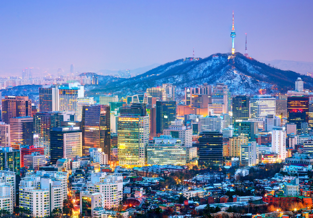 Cityscape of Seoul at dusk, with illuminated buildings in the foreground and Namsan Seoul Tower on a mountain in the background.