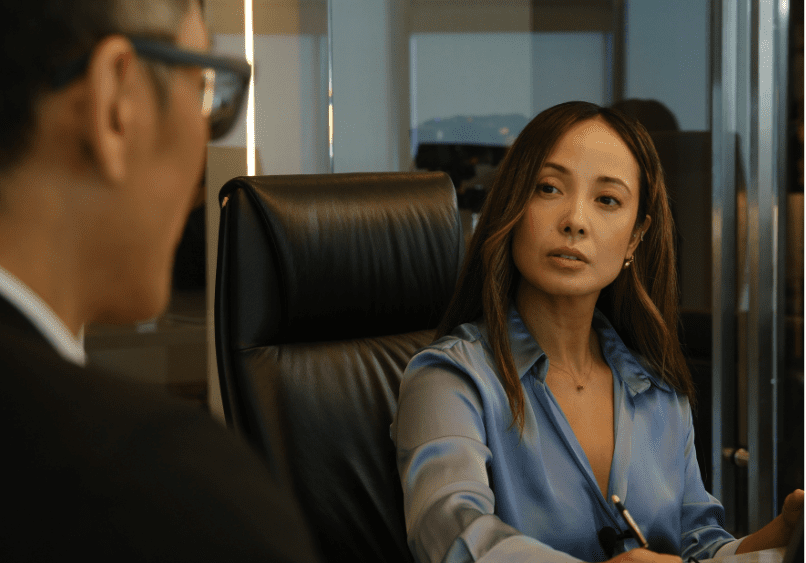 A woman in a blue blouse sits in a black chair, holding a pen and listening to a man in glasses during a meeting in an office setting.