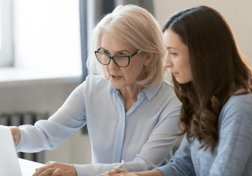 Two women sitting at a desk look at a laptop screen together; one points at the screen while the other listens attentively.