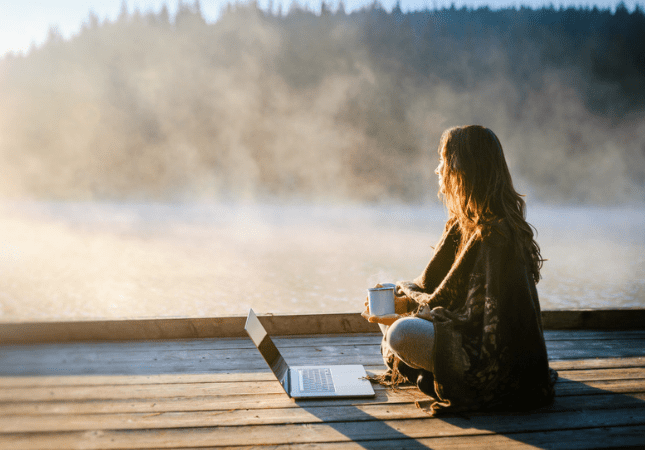 A person sits on a wooden deck by a misty lake, holding a mug and looking at the water, with an open laptop next to them.