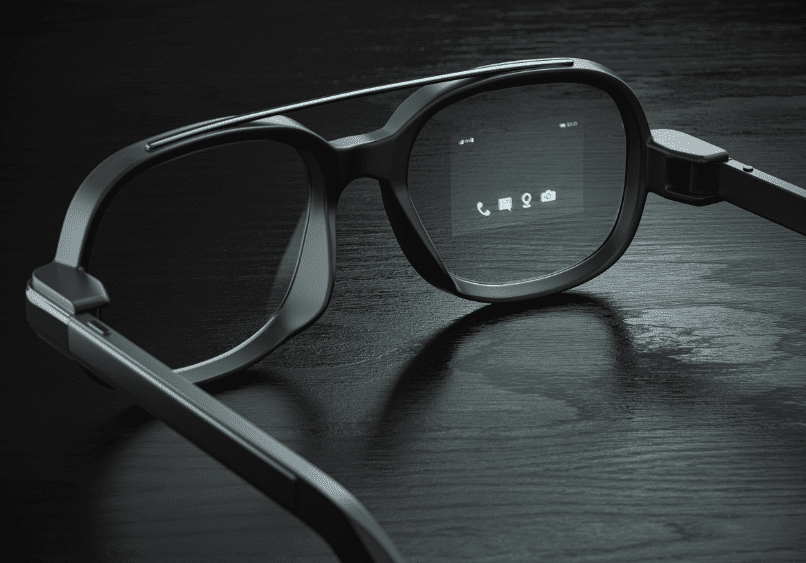 A pair of black smart glasses rests on a dark surface, displaying digital icons on the right lens.