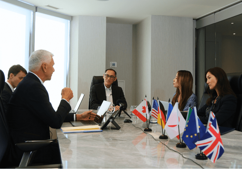 Five people sit around a conference table having a meeting, with six international flags displayed in the foreground.
