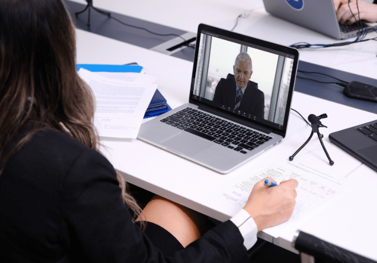 A person sits at a desk taking notes during a video call with a man displayed on a laptop screen.