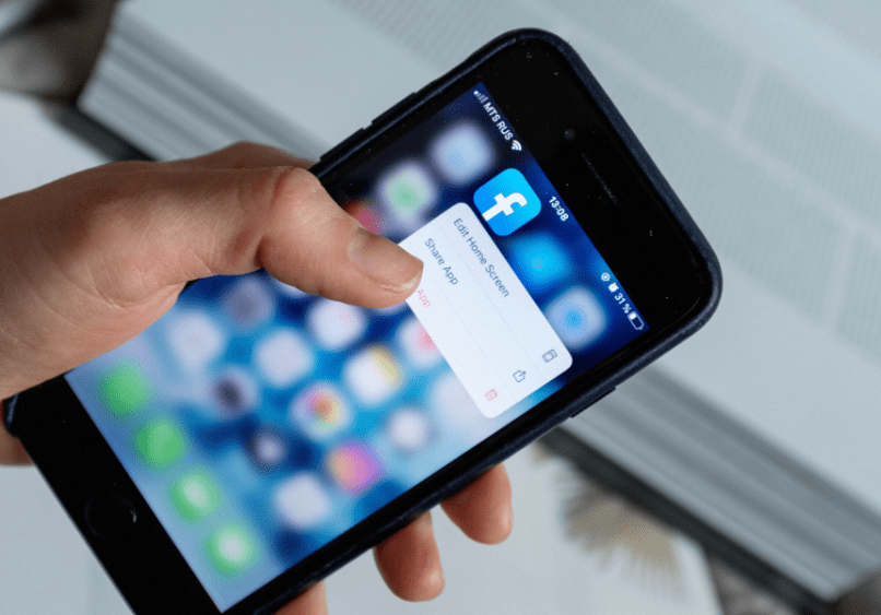 A hand holds a smartphone displaying the Facebook app, with a menu open offering options to edit the home screen or share the app.