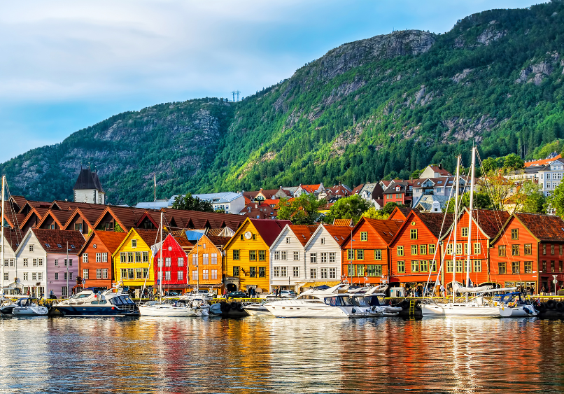 Row of colorful wooden buildings and boats along a waterfront, with green hills and scattered houses in the background under a partly cloudy sky.