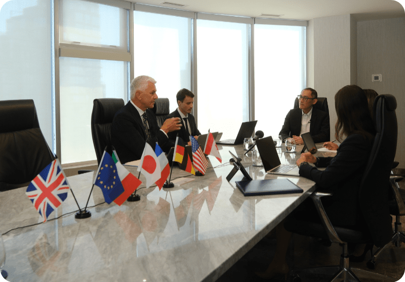 Four people in business attire have a meeting at a conference table with several international flags and laptops in front of them.