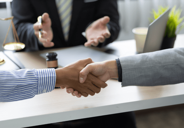 Two people shake hands across a desk in an office setting, with a third person gesturing in the background near legal documents and a gavel.