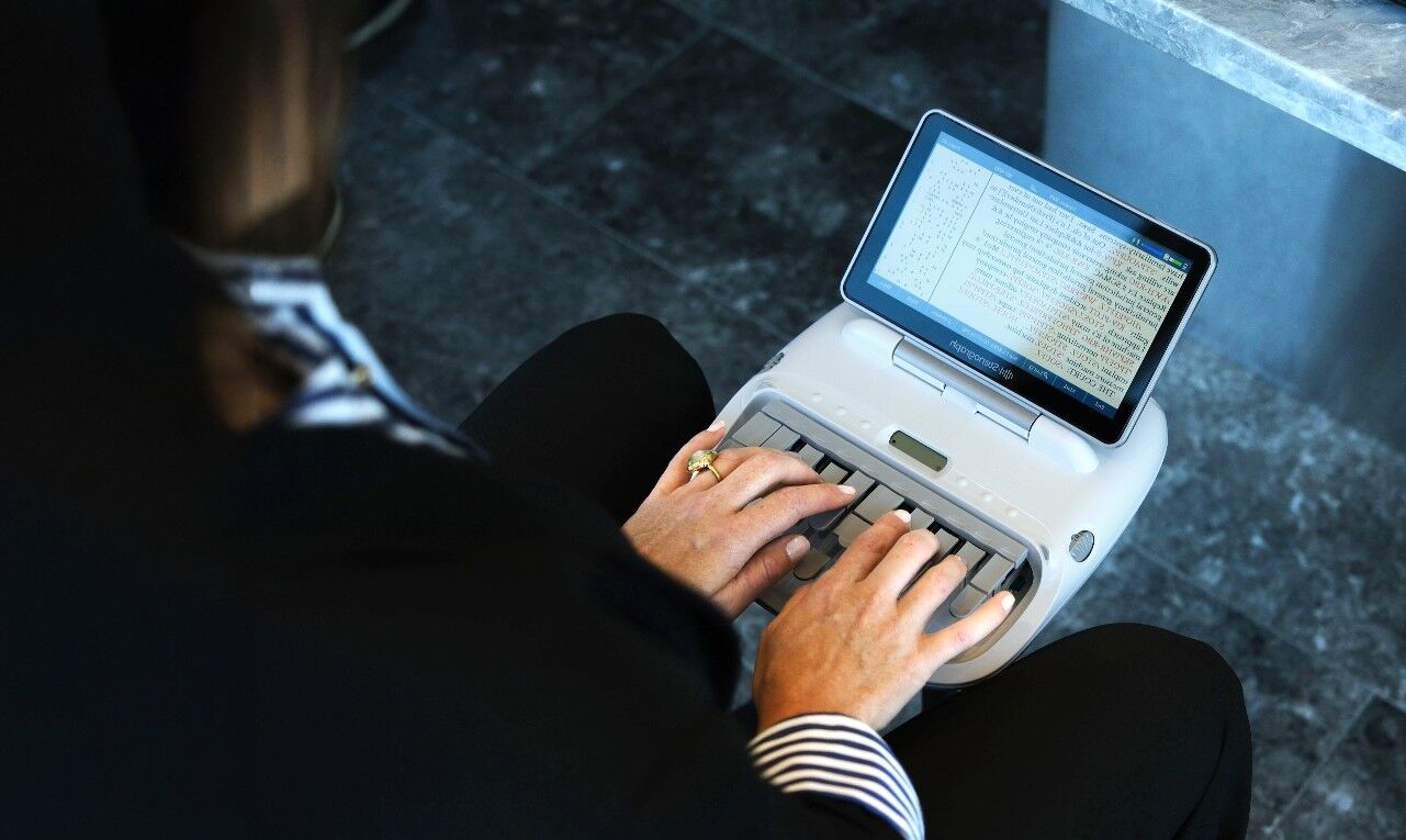 A person types on a stenotype machine connected to a tablet displaying text on the screen.