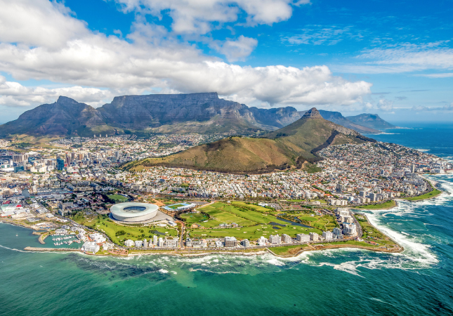 Aerial view of Cape Town, South Africa, showing Table Mountain, the city, Cape Town Stadium, and coastline with waves under a partly cloudy sky.